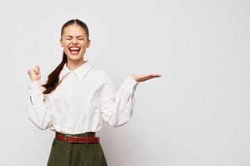 joyful woman, celebrating success with a big smile, wearing a white blouse and green pants, standing against a plain light grey background, conveying happiness and excitement