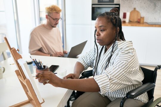 Two individuals engaging in creative work in home office setting, one painting on canvas while other works on laptop in background