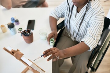 Person sitting in wheelchair, holding brush and painting canvas on an easel while focused on...