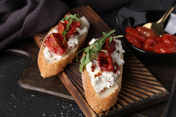 Wooden board of tasty tomato bruschetta with ricotta and arugula on black background, closeup