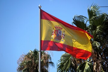Close Up der Flagge von Spanien, die stolz im Wind weht, mit Palmen und klarem blauen Himmel im...