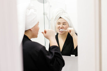 Woman with towel on head brushing teeth and looking at herself
