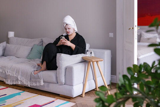 Plus size woman with face mask sitting on sofa and scrolling 