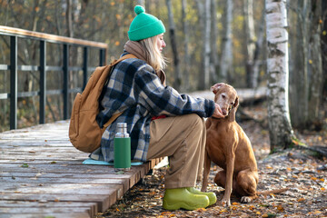 Woman traveler pauses hike, feeds Vizsla hunting dog by hand, sitting on eco trail in autumn nature...