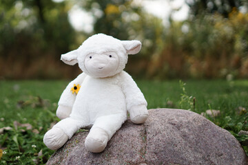 a cute plush lamb toy sitting on a rock in an outdoor setting, with a small yellow flower tucked...