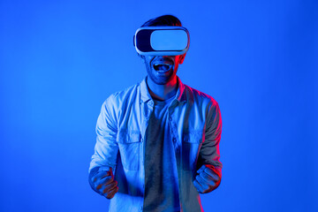Happy excited man watching at sport competition at virtual world while wearing VR headset at neon light background. Handsome teenager enter future innovation technology by using goggles. Deviation.