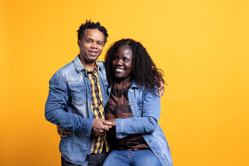 Fototapeta premium Romantic african american life partners embracing each other on camera, smiling and feeling cheerful against yellow background. Joyful pleased husband and wife hugging with affection.