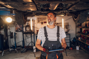 Happy auto mechanic worker at shop with tablet under chassis looking away