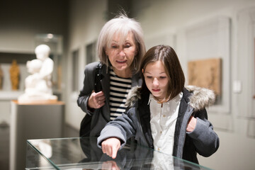 Positive senior woman and emotional inquisitive tween girl observing with interest arts and crafts...