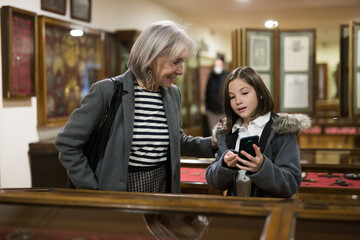 Cute curious tween girl browsing internet on smartphone in search of information about showpieces while visiting local history museum with her grandmother