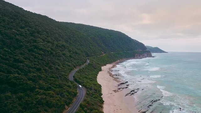 4K drone video of a camper van driving along a stunning long road next to a beautiful beach in Esperance, Western Australia. White waves from the blue ocean are crashing against the white sand beach.