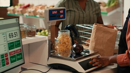 Merchant using scale at cash register checkout in organic shop, weighting freshly harvested fruits and veggies from local farm. African american seller preparing homegrown produce, eco friendly.