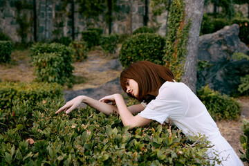 Elegant woman in white dress leaning against lush green bush in serene park garden setting
