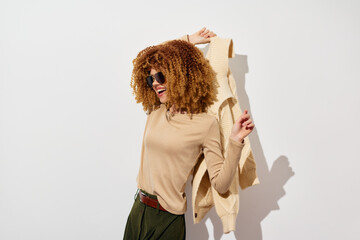 Fashionable woman with curly hair posing against a plain white background, wearing a beige outfit and sunglasses, showcasing a stylish look with confidence and joy
