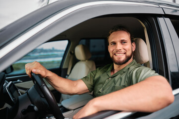 Smiling man in a green shirt sitting in a car, posing through the open driver s window, with a blurred urban background