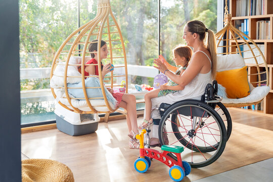 Mother in wheelchair playing with daughters in sunlit room.