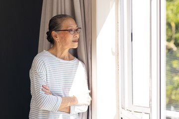 Looking out window, elderly asian woman in striped shirt enjoying sunlight indoors