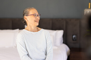 Smiling elderly Asian woman wearing glasses sitting on bed in cozy bedroom