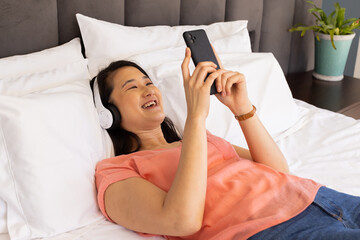 Listening to music with headphones, Asian woman smiling and using smartphone in bed