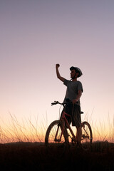 Fototapeta premium Mountain biker takes a break at dusk, raising his arm in victory. The scenic sunset and peaceful atmosphere capture a sense of accomplishment and a passion for outdoor adventure.
