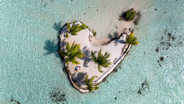Happy couple sit together on remote, romantic tropical island shaped like a heart. Aerial view captured by drone. 
