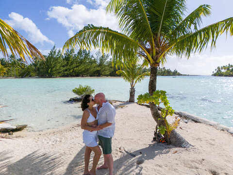Man and woman kissing while on a small private island with a tiny beach covered with sand and a palm tree. The romantic location is surrounded by beautiful ocean sea water in French Polynesia. 