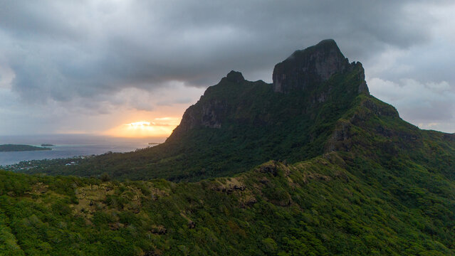 Aerial view captured by drone of Mount Otemanu on the tropical island Bora Bora in French Polynesia. The sun is setting under low dark clouds behind the volcano. 