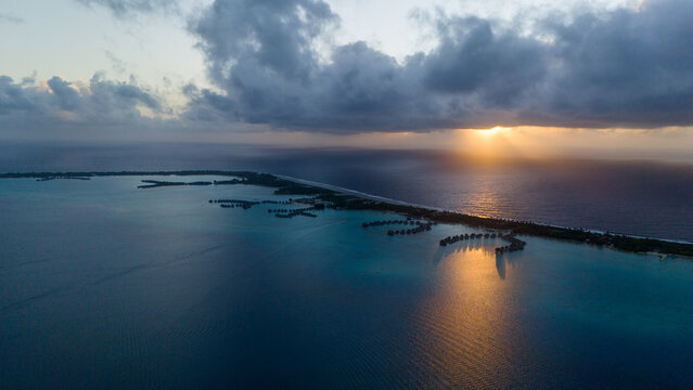 Aerial view captured by drone overlooking tropical island resorts with overwater bungalows on Bora Bora motu in French Polynesia. Azure and turquoise colored ocean sea water. Low clouds above sunrise.