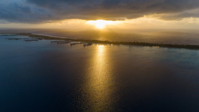 Aerial view captured by drone overlooking tropical island resorts with overwater bungalows on Bora Bora motu in French Polynesia. Azure and turquoise colored ocean sea water. Low clouds above sunrise.