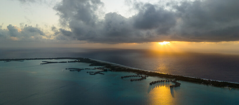 Aerial view captured by drone of overwater bungalows near the tropical vacation island of Bora Bora in French Polynesia. The early morning sun is rising in the background.