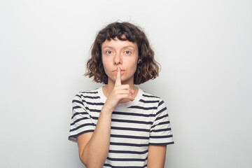 Curly-haired woman making shushing gesture against white wall