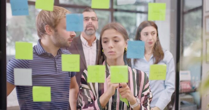 Work colleagues discussing startup project ideas. Team of caucasian business people standing near glass window with sticky notes. Scrum methodology with colorful stickers.