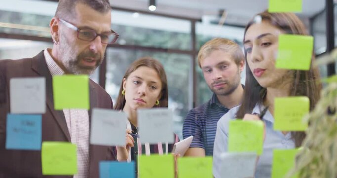 Work colleagues discussing startup project ideas. Team of caucasian business people standing near glass wall with sticky notes. Scrum methodology with colorful stickers. Forward camera movement.