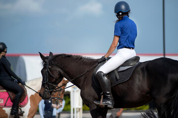 Backside, teenage equestrian girl in protective helmet sits astride a horse waiting for practice...