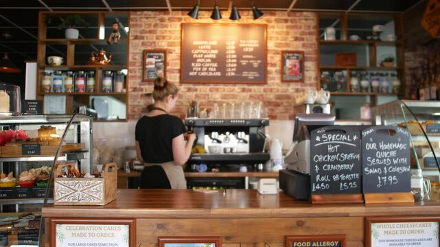 A female barista making coffee at a cafe counter with baked goods on display and chalkboard menus listing specials. The cozy setting features warm brick walls and wooden accents