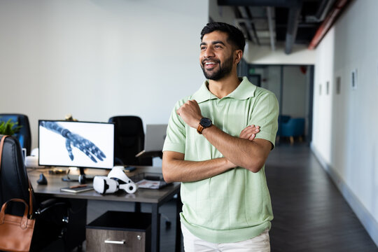 Smiling Indian man in office with robotic arm on computer screen in background