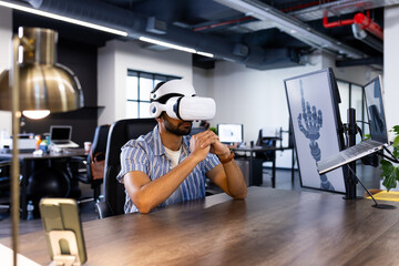 Using VR headset, Indian man sitting at desk in modern office with robotic hand display