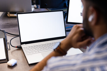 Using laptop with blank screen, Indian man thinking and wearing wireless earbuds, copy space