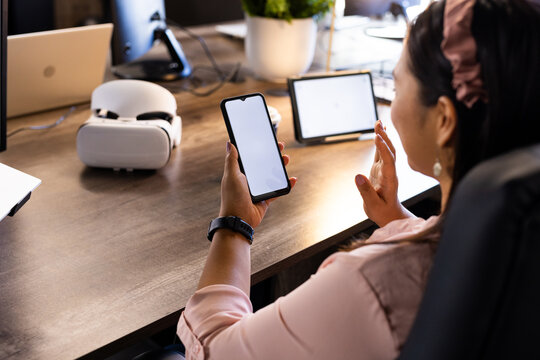 Using smartphone, woman in office waving at tablet with VR headset nearby, copy space
