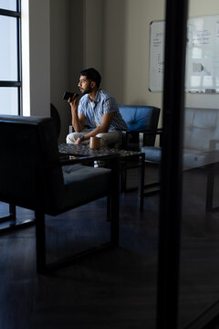 Holding smartphone, Indian man sitting in modern office with coffee cup, copy space