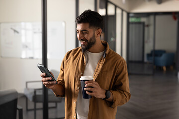 Holding coffee cup, Indian man smiling and checking smartphone in modern office