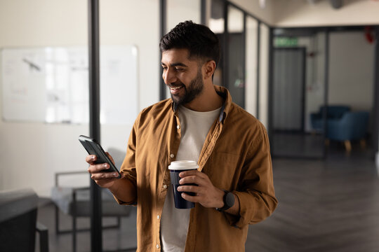 Holding coffee cup, Indian man smiling and checking smartphone in modern office