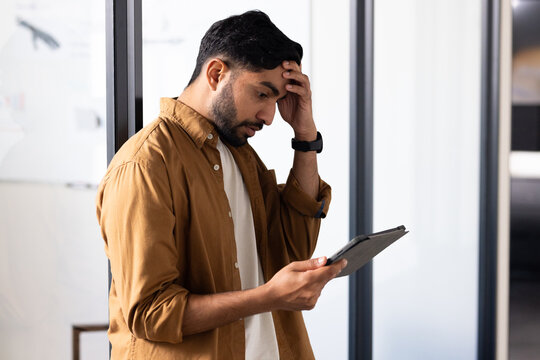 Worried Indian man holding tablet, reading important information in modern office
