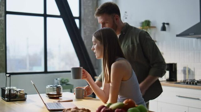 Husband pouring coffee wife cup looking laptop at home kitchen. Smiling couple
