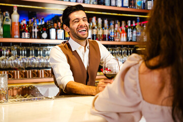 Caucasian professional bartender or mixologist making a cocktail for women at a bar. An attractive barman served a glass of wine with a smile to a woman. Bartender service at the night club restaurant