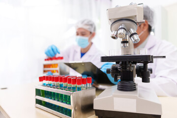 Two Asian scientists in lab coats sit at a table in a laboratory, examining samples through a microscope. The room is equipped with scientific tools, liquids, and chemistry materials.