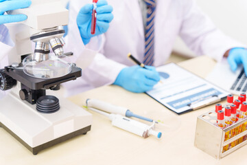 Two Asian scientists in lab coats sit at a table in a laboratory, examining samples through a microscope. The room is equipped with scientific tools, liquids, and chemistry materials.