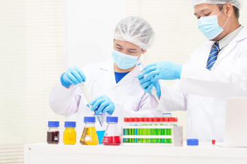 Two Asian scientists in lab coats sit at a table in a laboratory, examining samples through a microscope. The room is equipped with scientific tools, liquids, and chemistry materials.