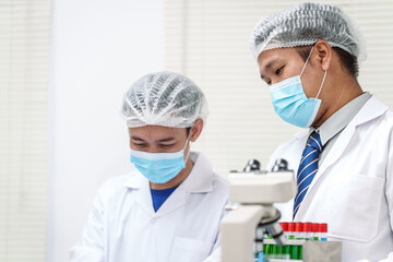 Two Asian scientists in lab coats sit at a table in a laboratory, examining samples through a...