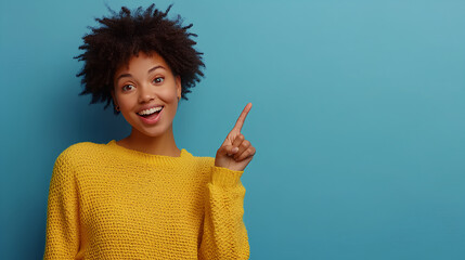 Cheerful Afro woman points away on copy space, discusses amazing promo, gives way or direction, wears yellow warm sweater, has pleasant smile, feels optimistic, isolated over blue background